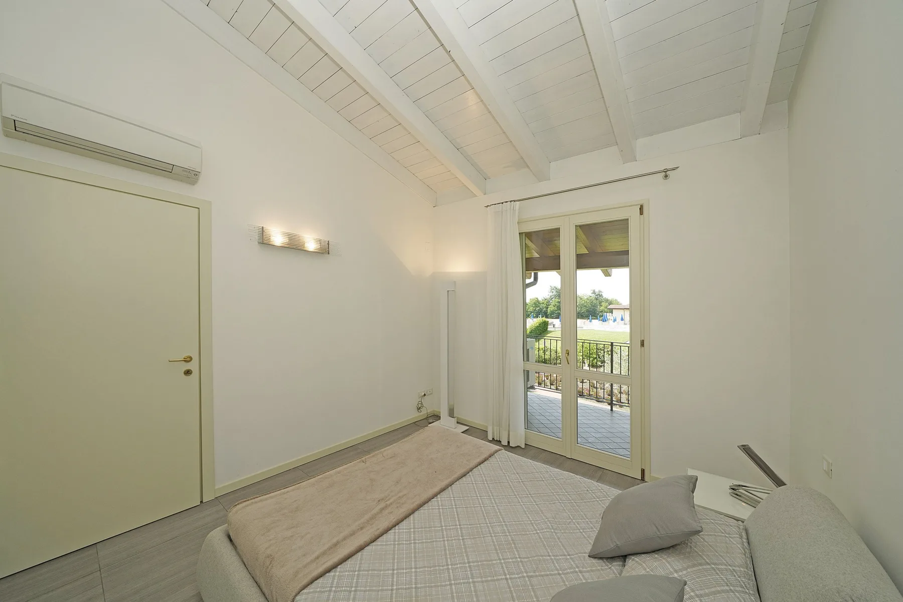 Un dormitorio luminoso con techo blanco inclinado y puertas de vidrio que dan a un balcón.