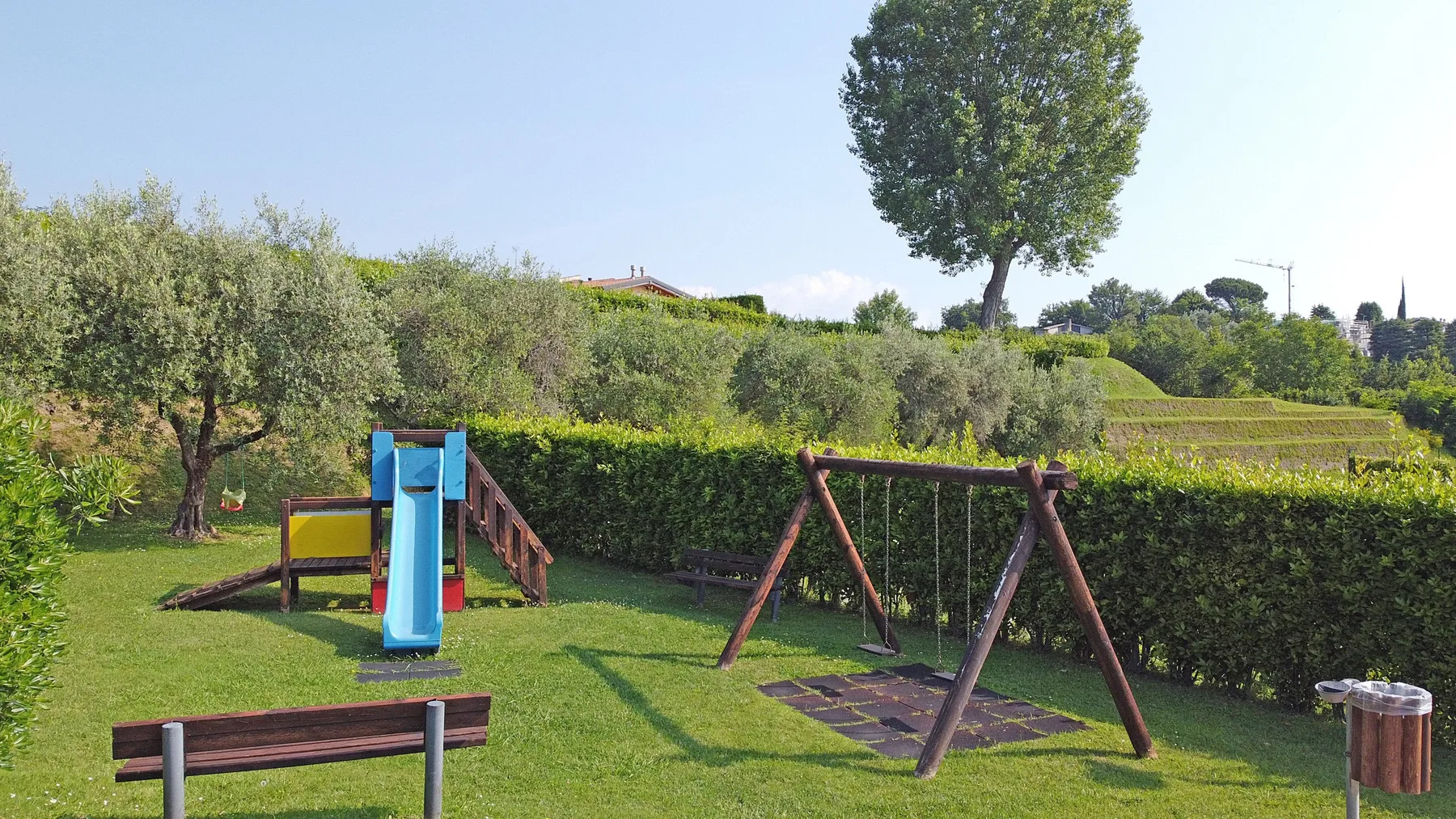 Una zona de juegos en un jardín con césped tiene un tobogán, columpios y un banco de madera.