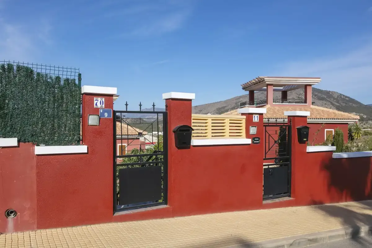 A red-walled property entrance with metal gates and a mountain in the background.