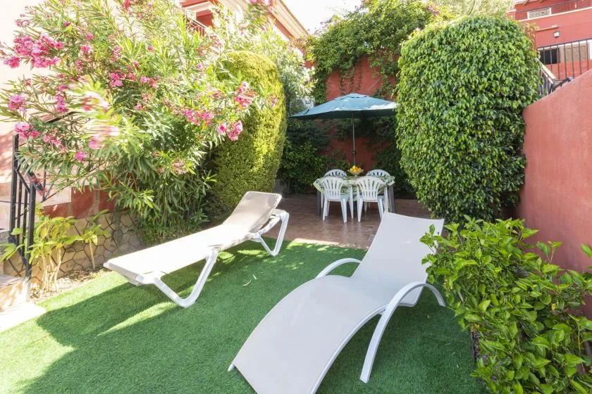 A small garden patio has white lounge chairs, a dining set, and flowering greenery.