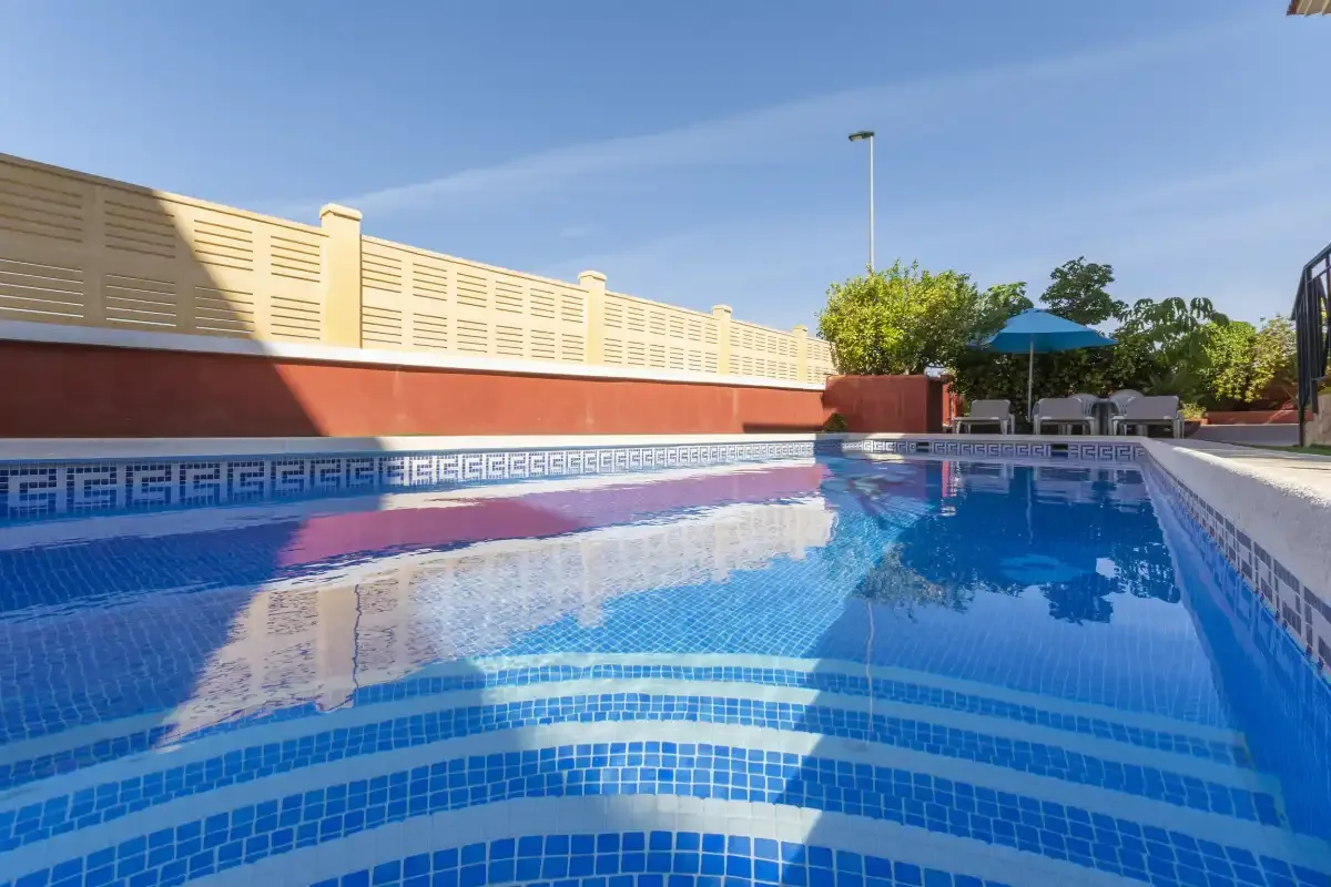 An outdoor swimming pool with blue tile and a shaded seating area.