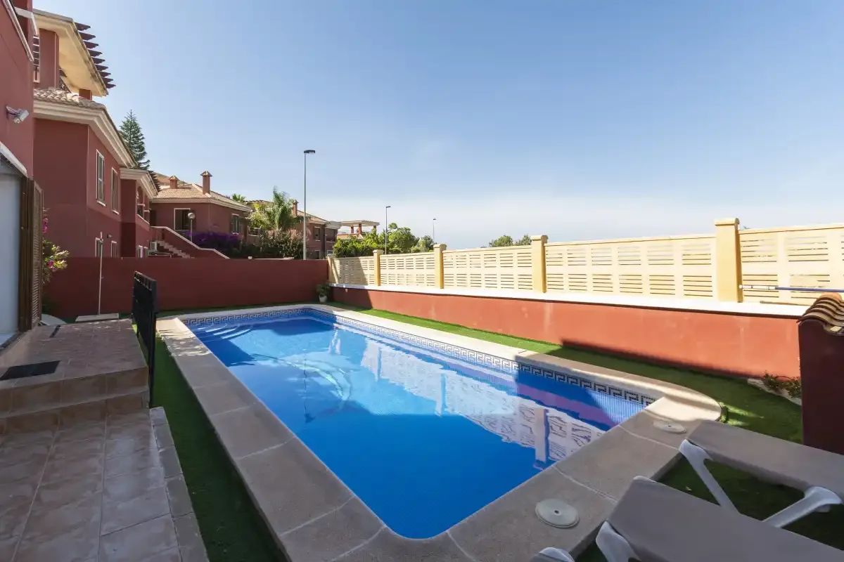 A swimming pool sits beside a red house and a pale perimeter fence.