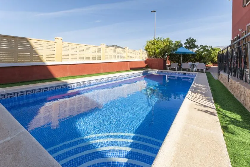 An outdoor swimming pool beside a patio with seating and a blue umbrella.