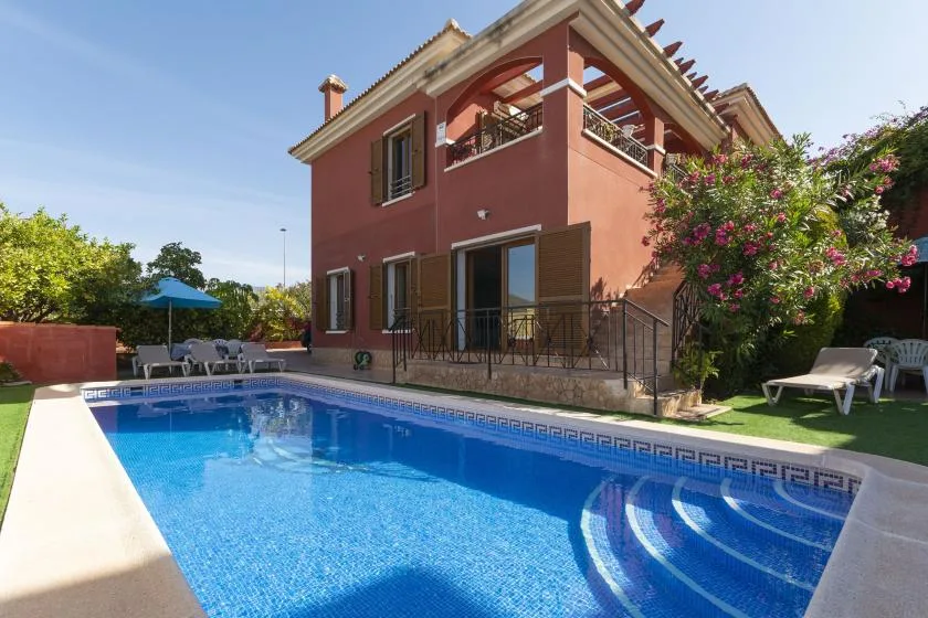 A swimming pool beside a red house, with lounge seating and flowering shrubs.