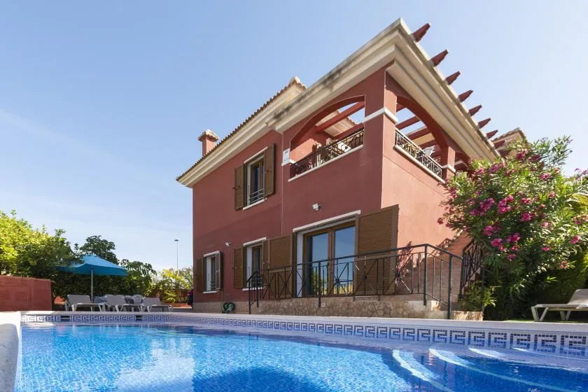 A red two-story house overlooks a blue swimming pool in a sunny garden.