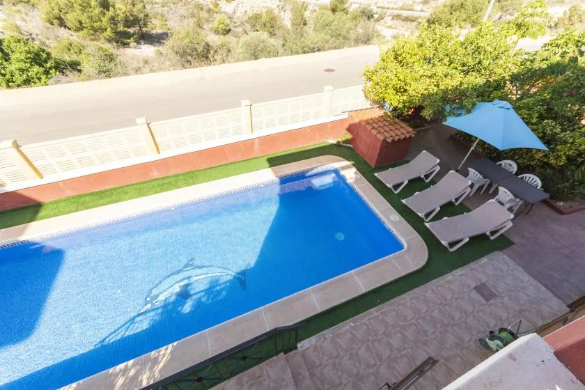 An outdoor swimming pool sits beside a paved patio with lounge seating and an umbrella.