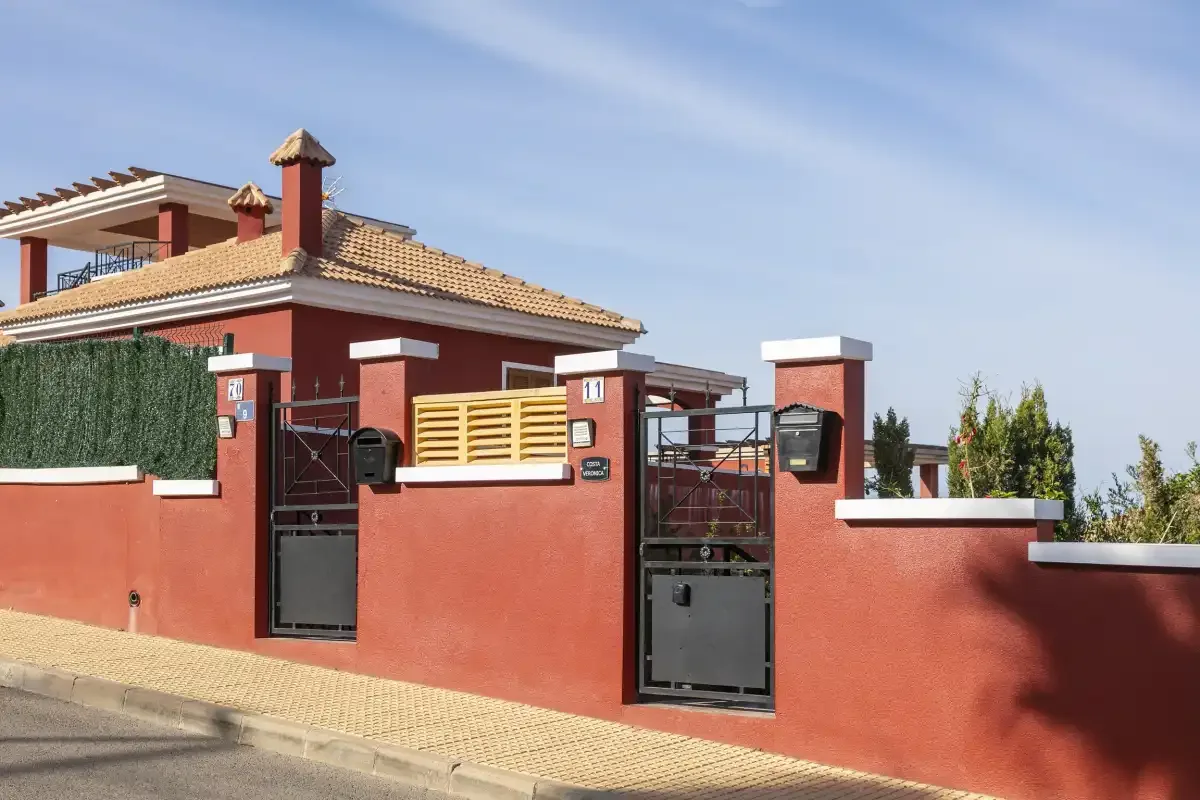 A red stucco house stands behind a matching boundary wall and gate.