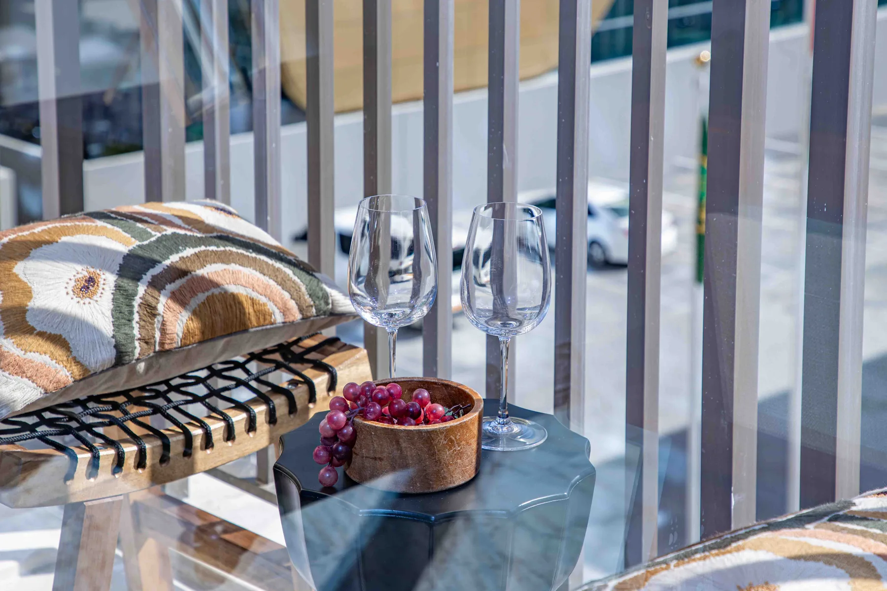 Zona de estar en un balcón con mesa de vidrio, copas de vino y uvas.