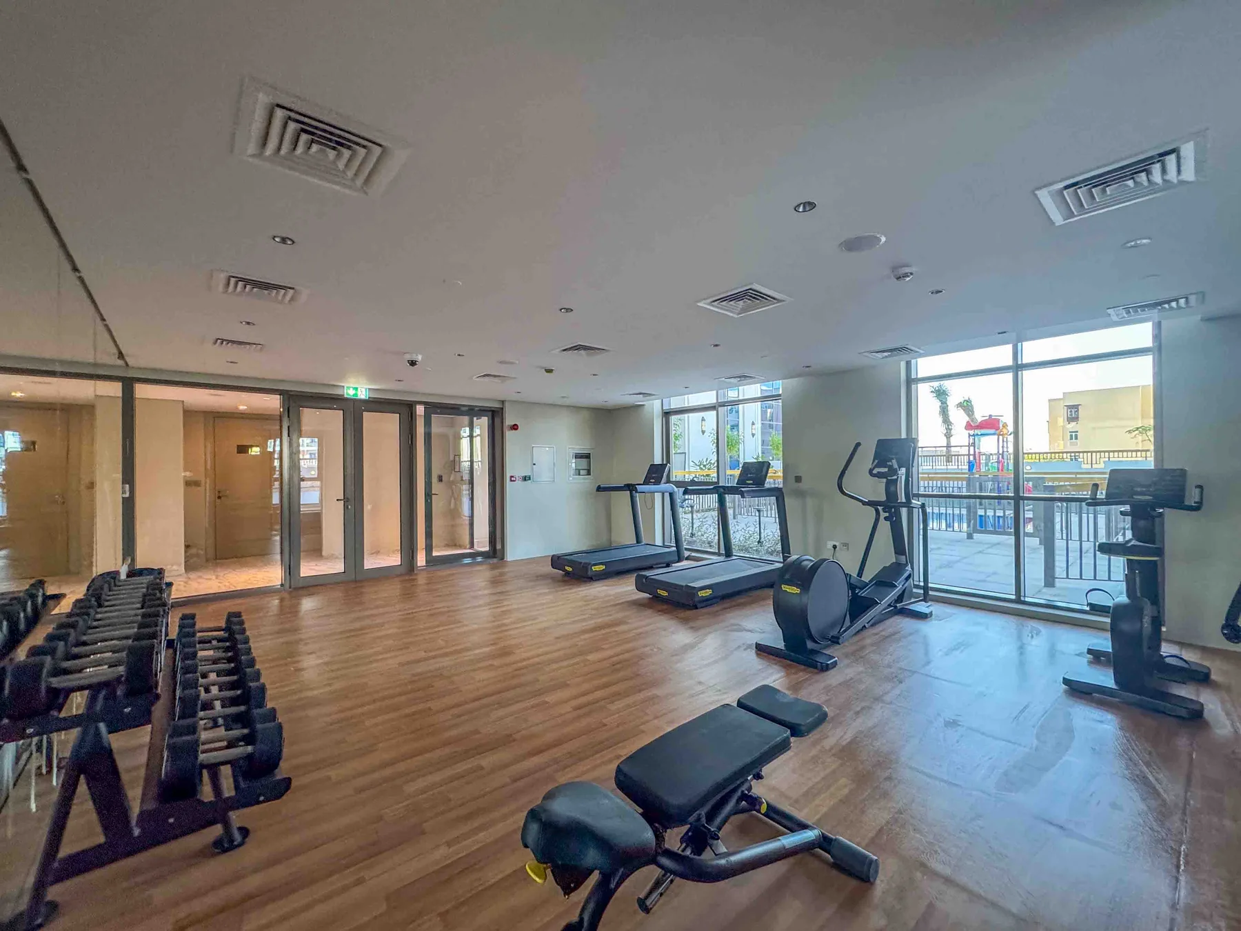 Un gimnasio con bicicletas estáticas, cintas de correr y pesas junto a grandes ventanas.