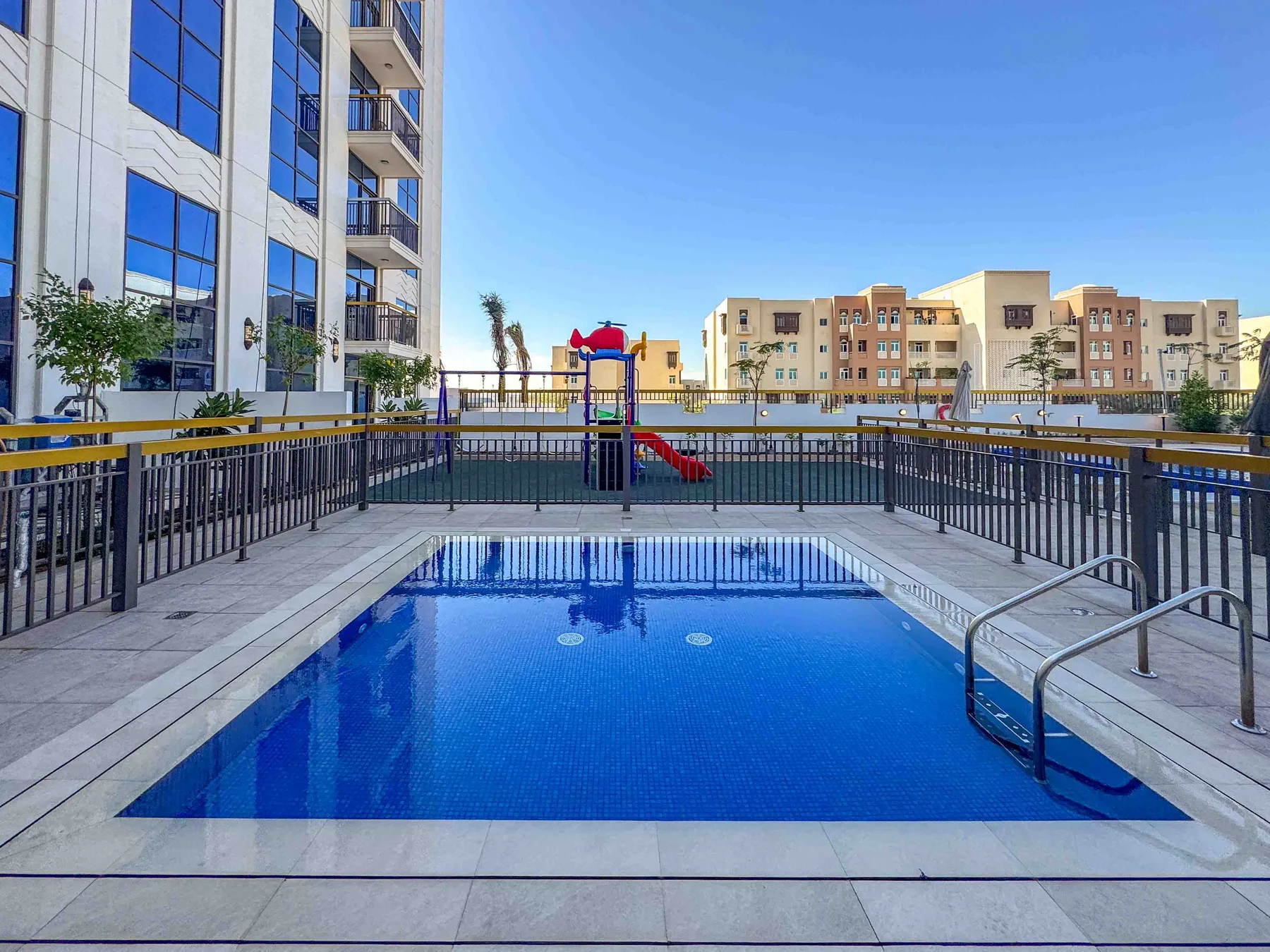 Una piscina azul está junto a un área de juegos cercada entre edificios de apartamentos.