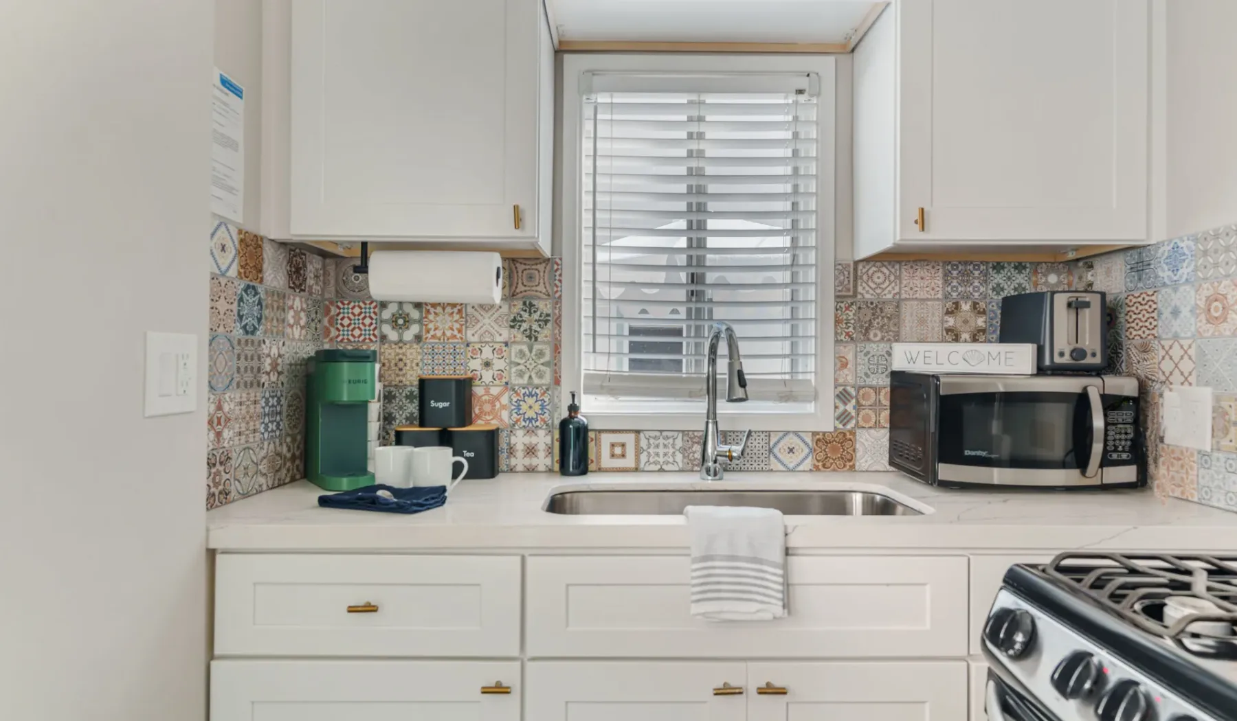 Una cocina luminosa con gabinetes blancos, azulejos estampados y una ventana sobre el fregadero.