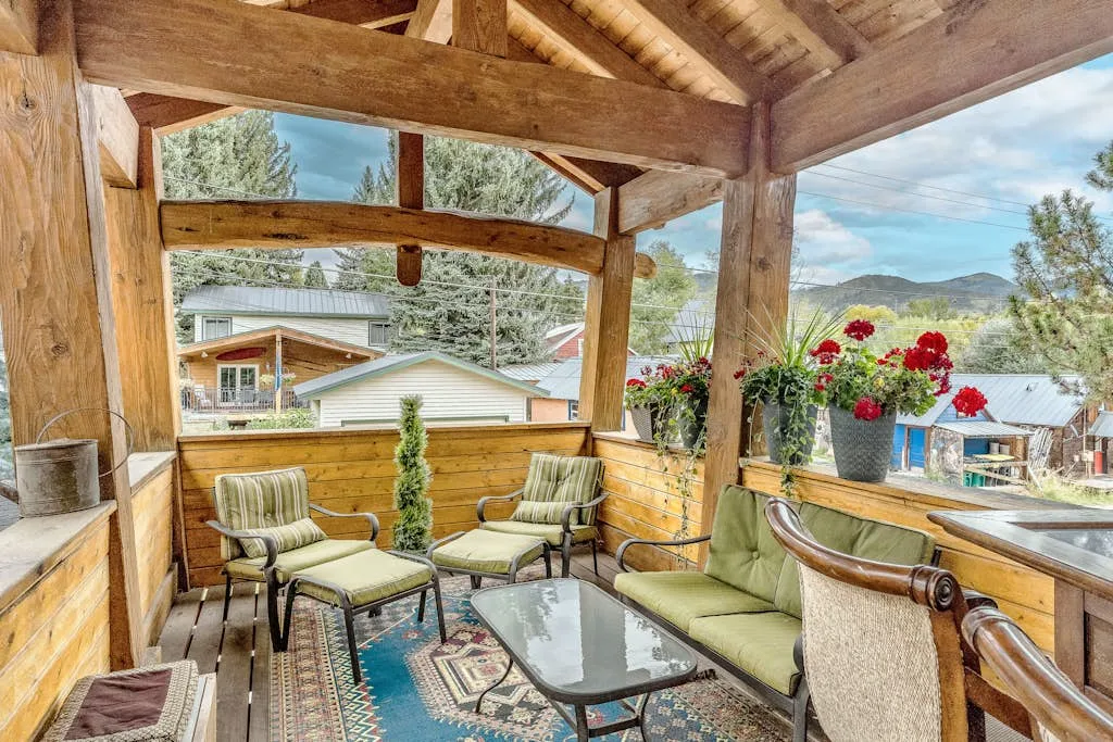 Un patio cubierto de madera tiene asientos acolchados y vistas de casas y árboles cercanos.