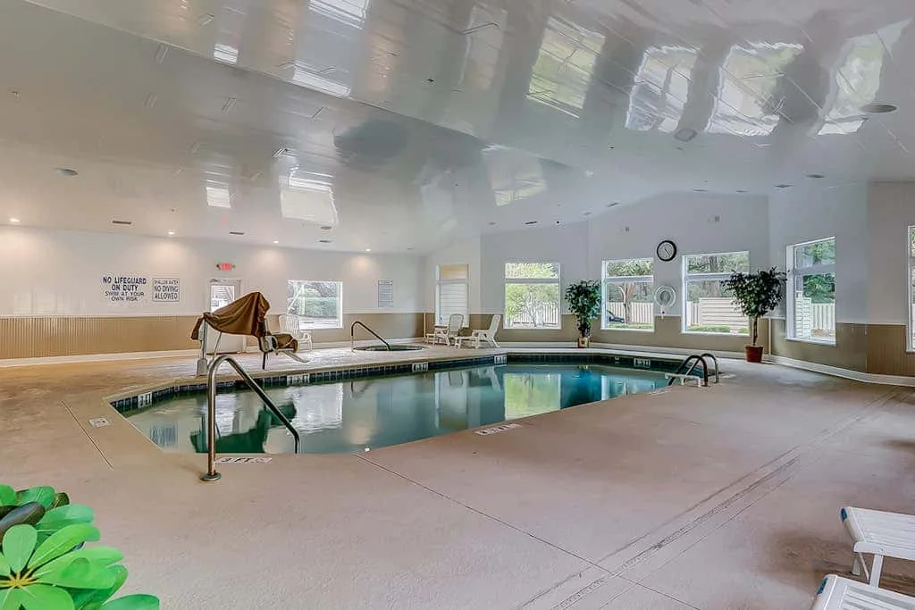 An indoor swimming pool with a tiled deck, windows, and seating.