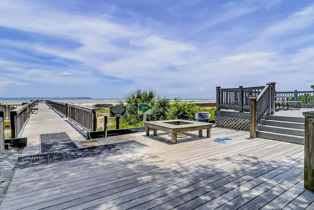 A wooden boardwalk deck with railings, steps, and coastal grasses under a cloudy sky.