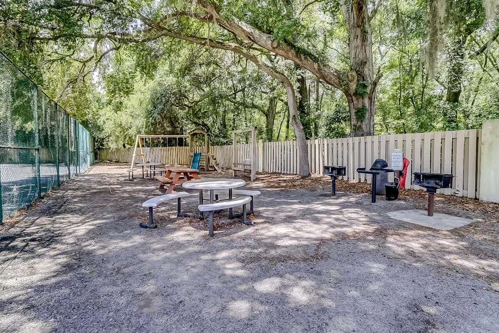 An outdoor picnic area with tables, grills, and a fenced play structure beneath trees.