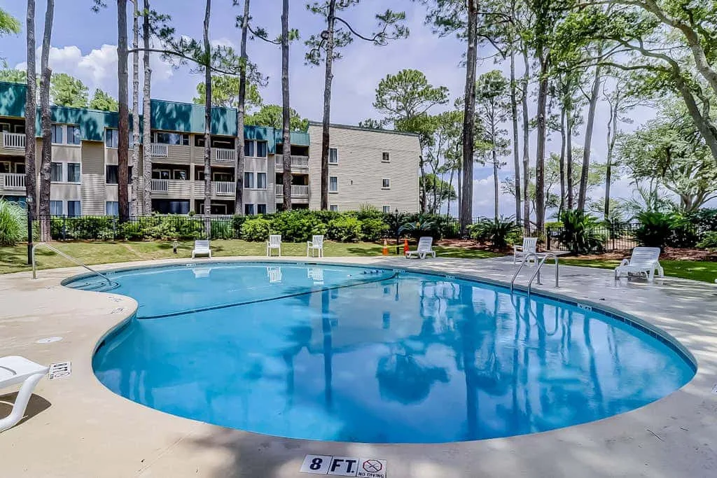 An outdoor swimming pool sits beside a multi-story building and tall trees.