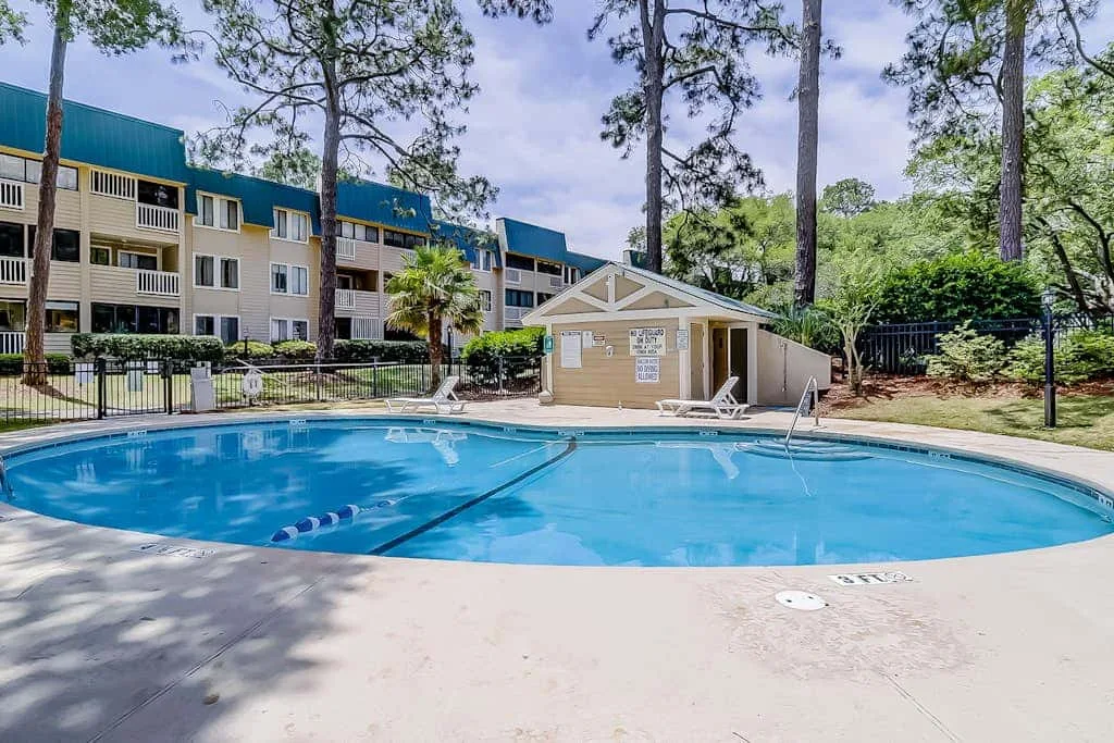 A swimming pool sits in front of a multi-story building and surrounding trees.