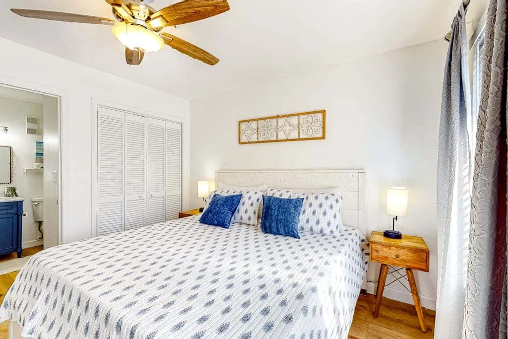 A bright bedroom with a patterned bedspread, blue pillows, and a ceiling fan.
