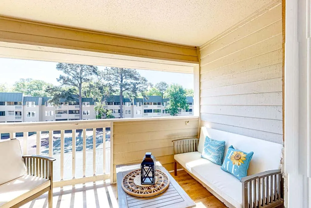 A covered balcony has seating, a small table, and views of nearby buildings and trees.