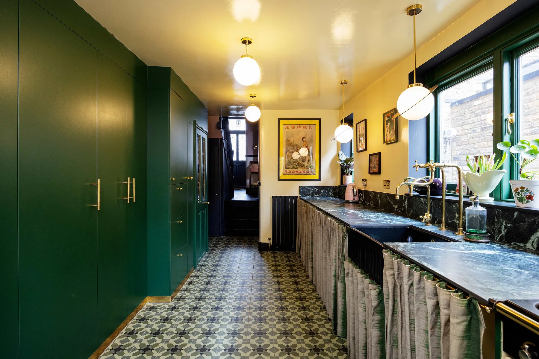 Una cocina estrecha con gabinetes verde oscuro, suelo estampado y una encimera larga bajo las ventanas.
