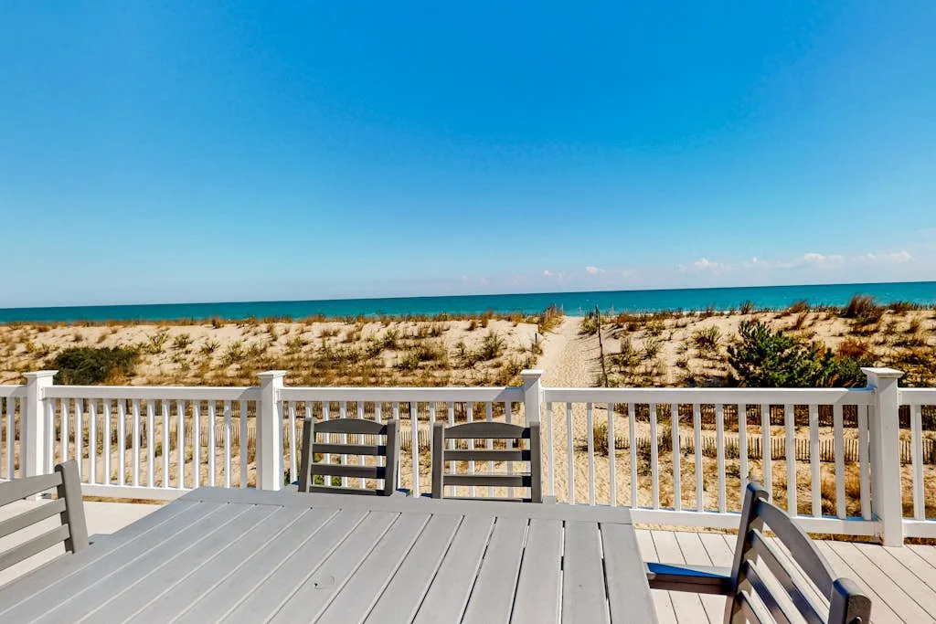 A wooden deck overlooks sandy dunes and the ocean under a clear sky.