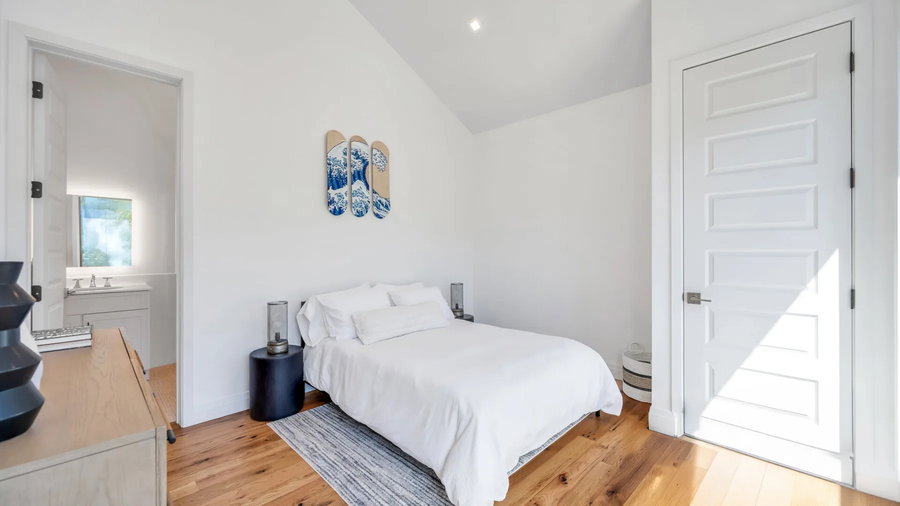 A bright bedroom with a bed, wood flooring, and an open doorway to a bathroom.