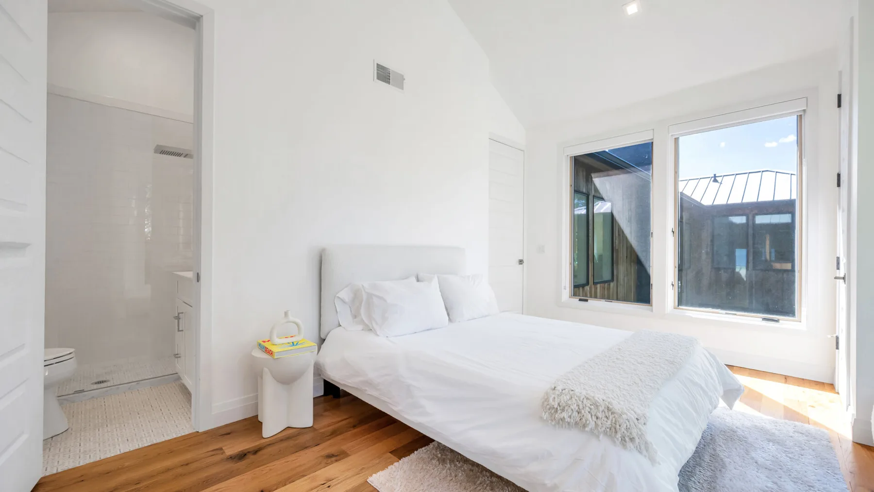 A bright bedroom with a white bed beside large windows.