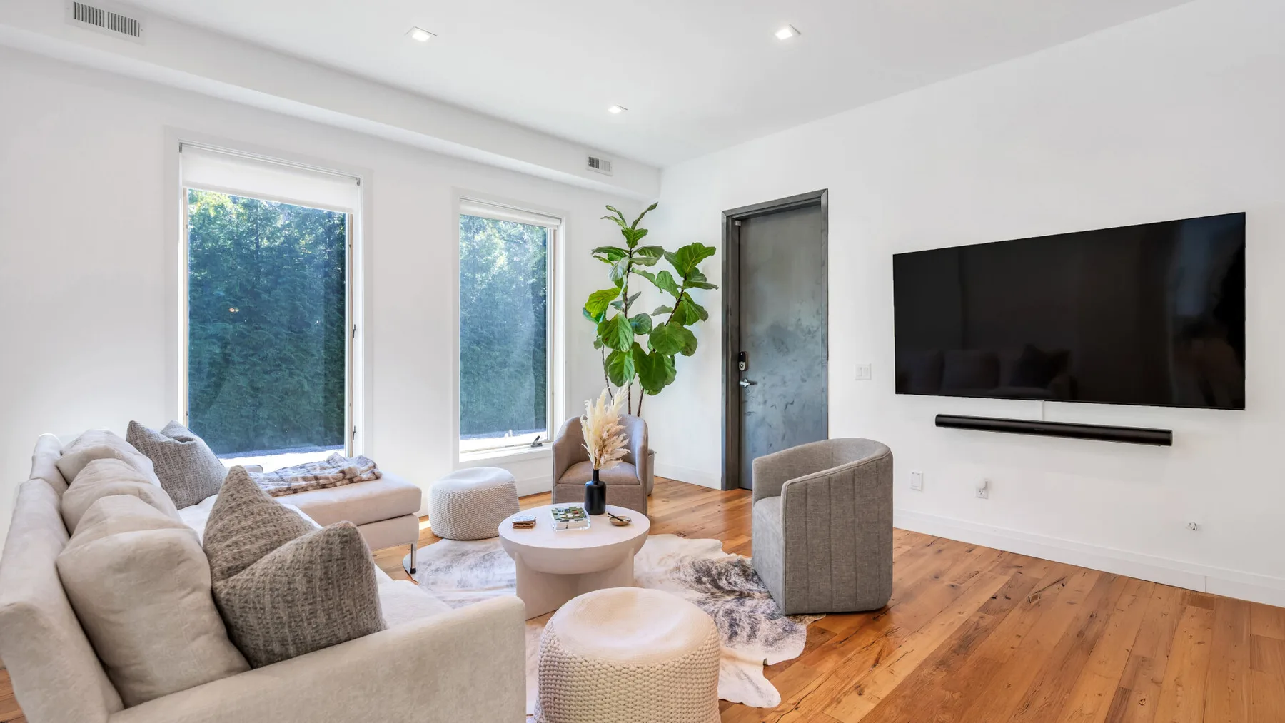 A bright living room with seating, a wall-mounted television, and large windows.