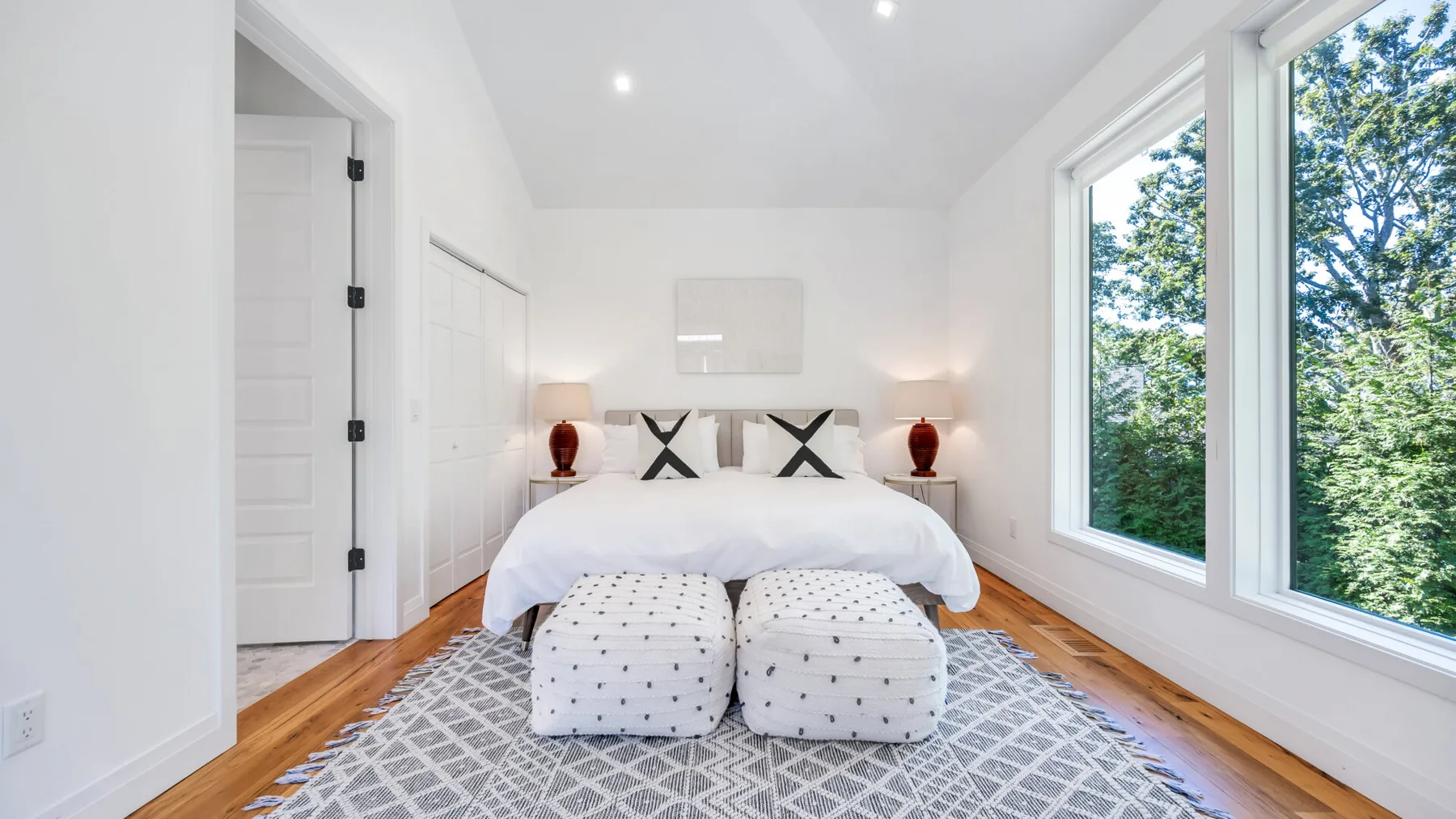 A bright bedroom with a bed, patterned rug, and large windows overlooking trees.