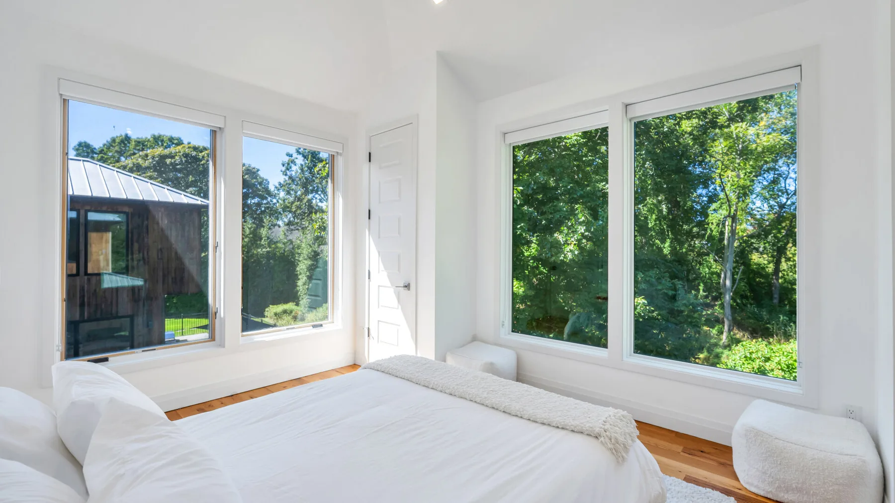 A bright bedroom with a bed beside large windows overlooking leafy trees.