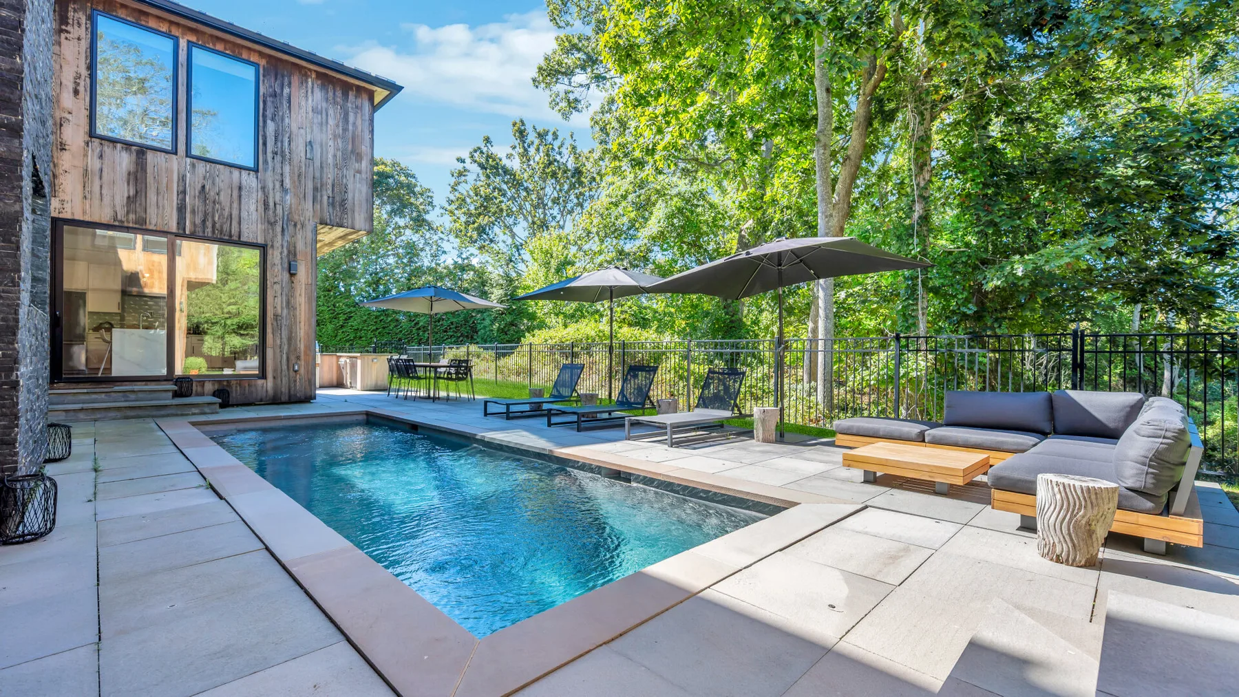 An outdoor pool beside a wooden-sided building, with seating and umbrellas on the patio.