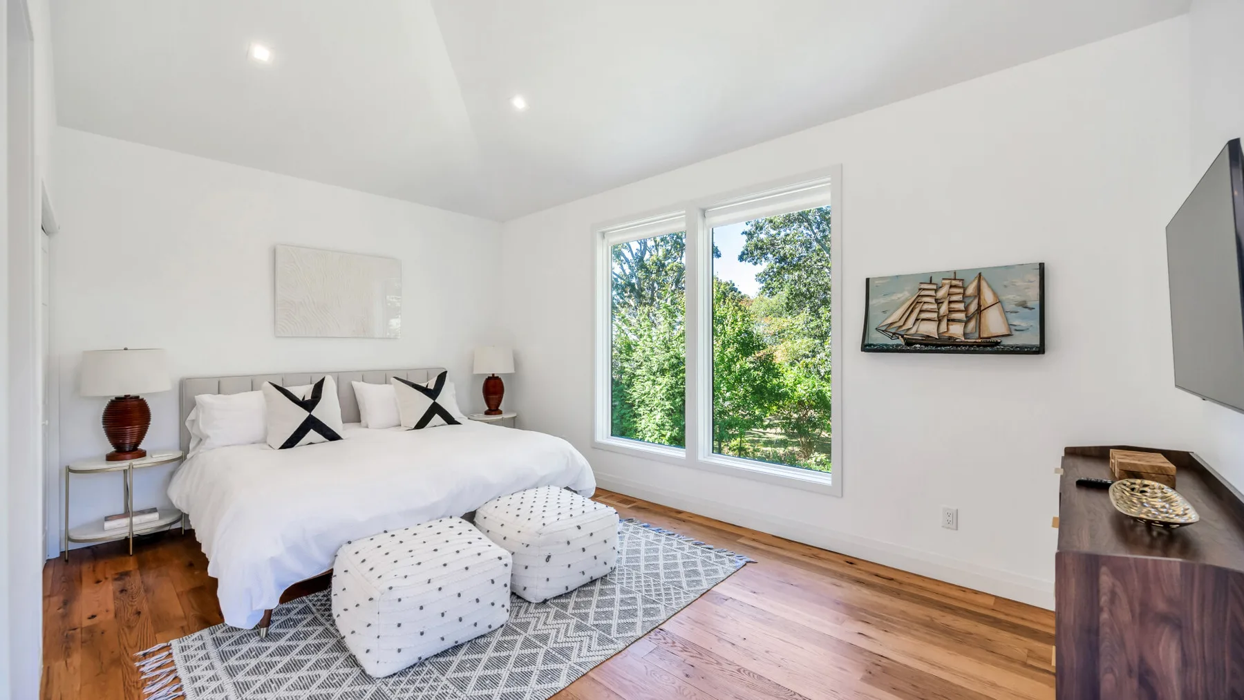 A bright bedroom with a bed, patterned rug, and large windows overlooking greenery.