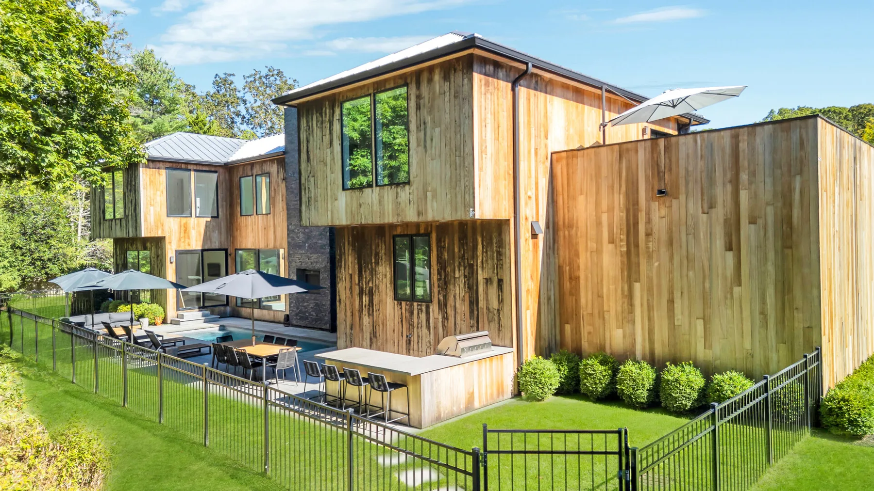 A modern wood-clad house stands beside a fenced patio with outdoor seating.