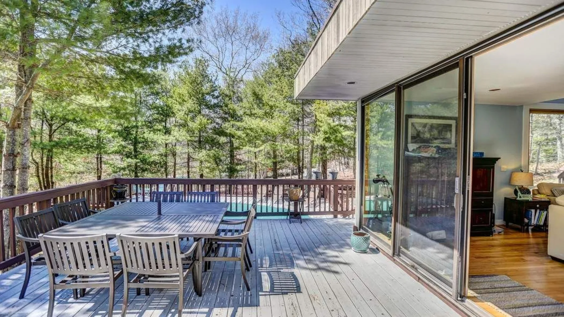 A wooden deck with outdoor seating overlooks a wooded area beside a glass-walled room.
