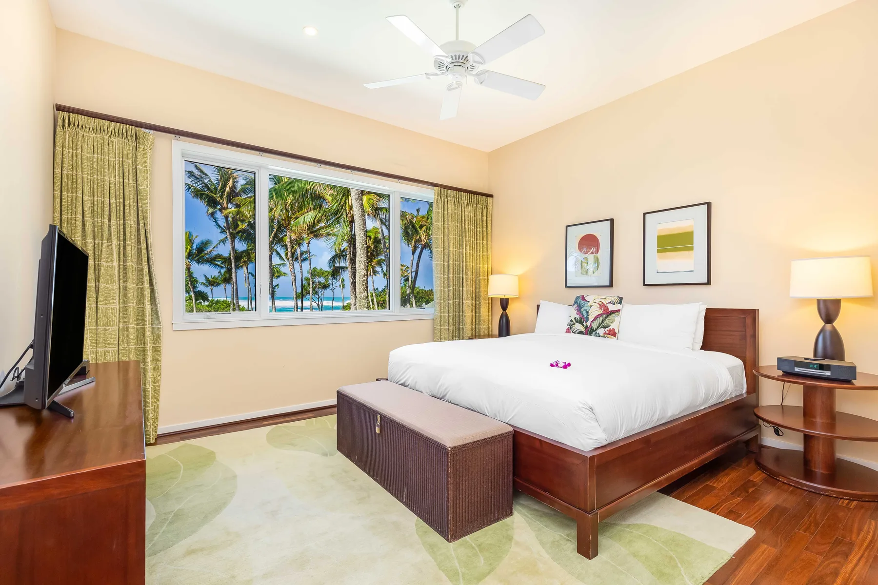 Un dormitorio luminoso con una cama grande, lámparas de noche y una ventana con vista a palmeras.