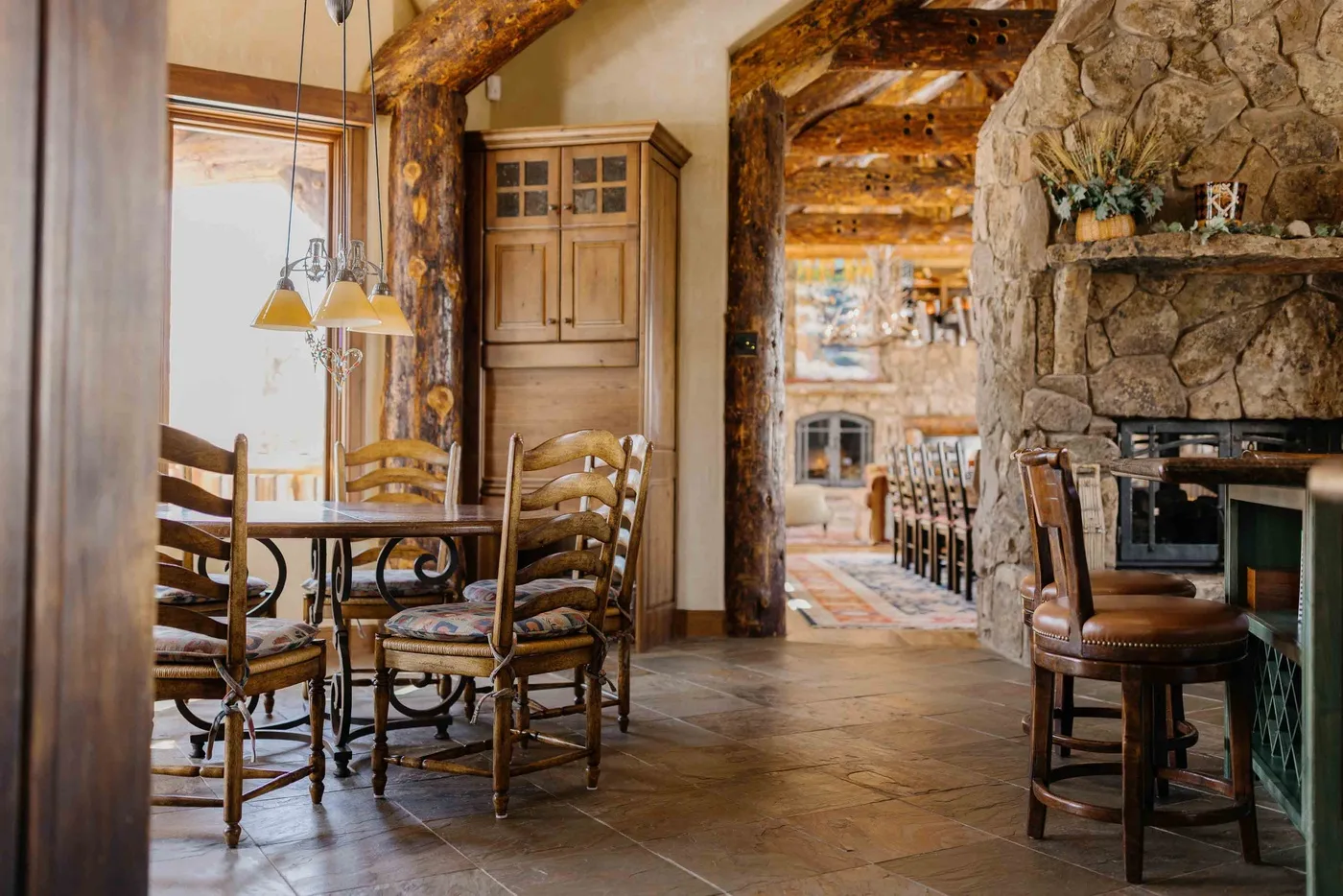 A rustic dining area with wooden seating beside a large stone fireplace.
