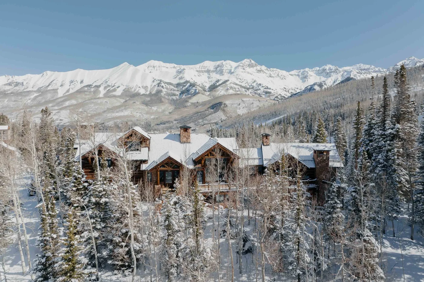 A lodge sits among snow-covered trees beneath a range of snowy mountains.