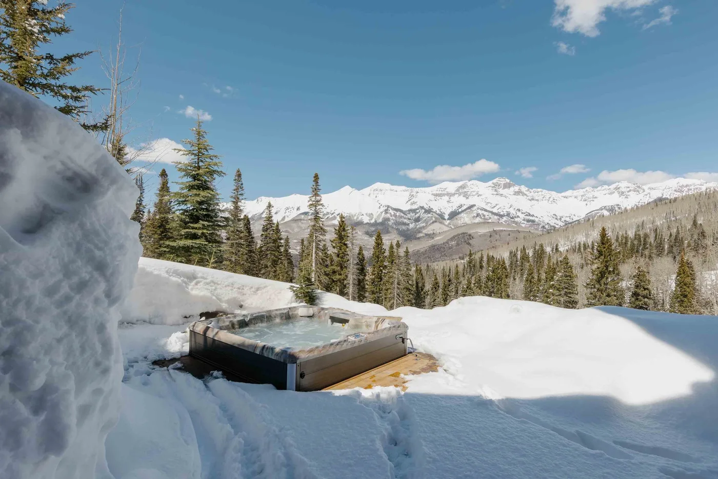 An outdoor hot tub sits in a snowy landscape with evergreen trees and mountains.