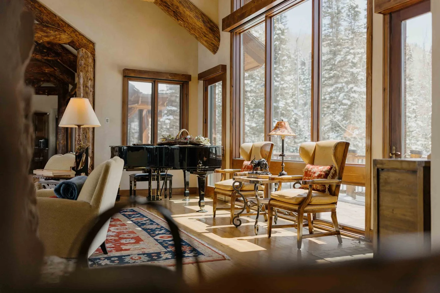 A sunlit living room with seating, a piano, and large windows overlooking trees.