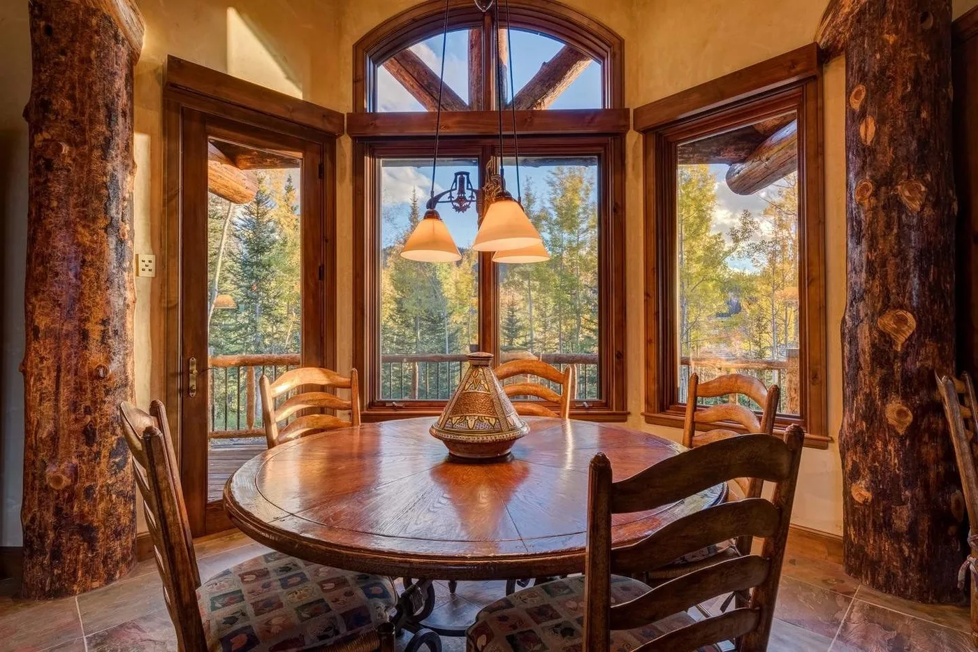 A dining area with a round wooden table beside large windows overlooking trees.