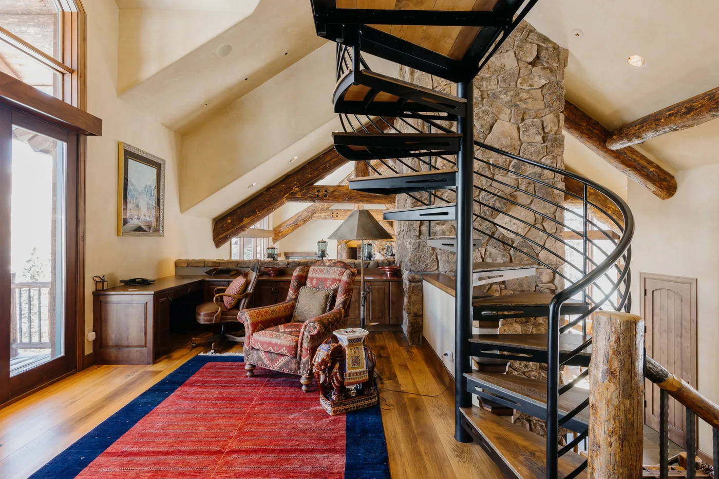 A rustic loft living area with patterned seating and a spiral staircase beside a stone column.