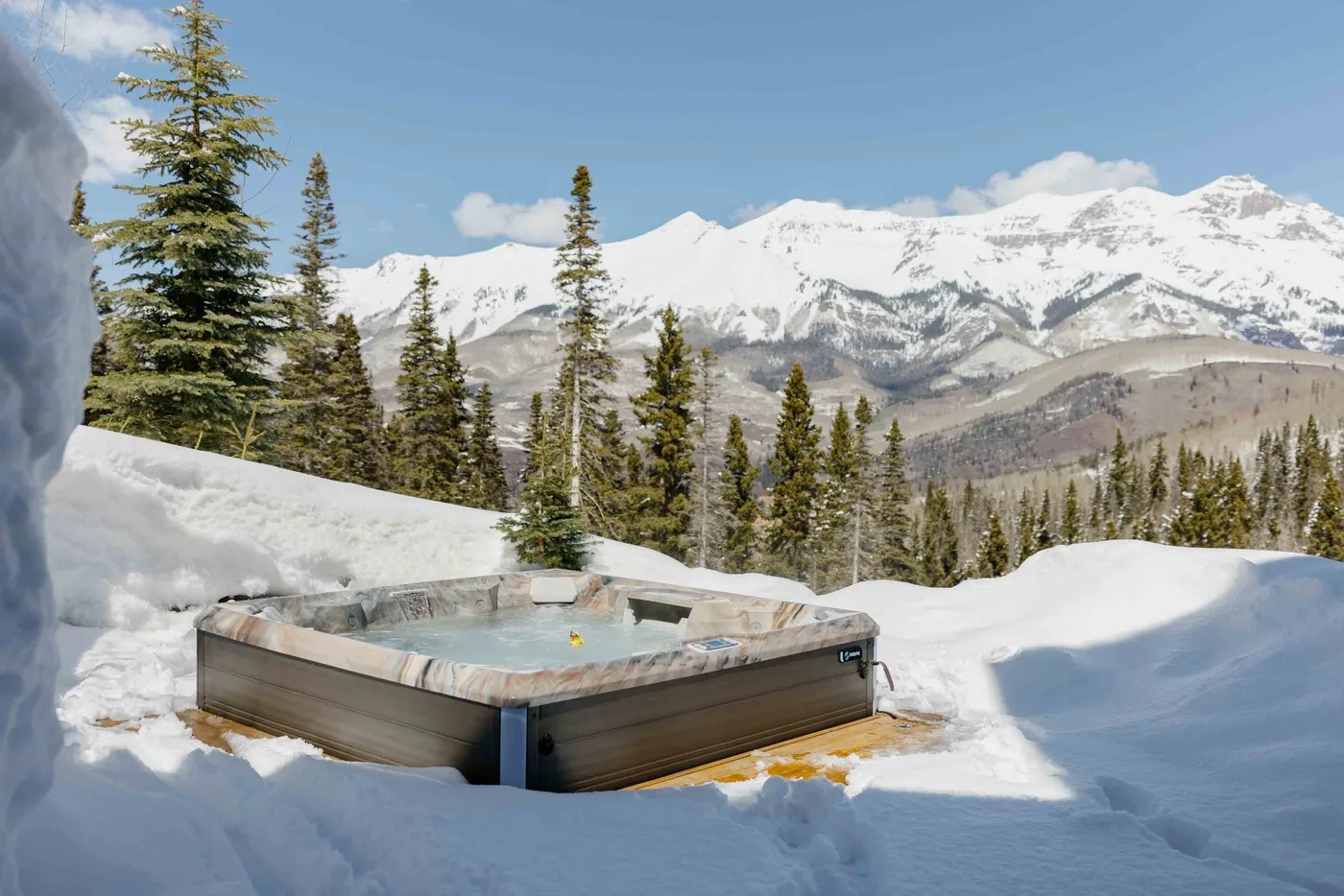An outdoor hot tub sits in a snowy mountain landscape among evergreen trees.