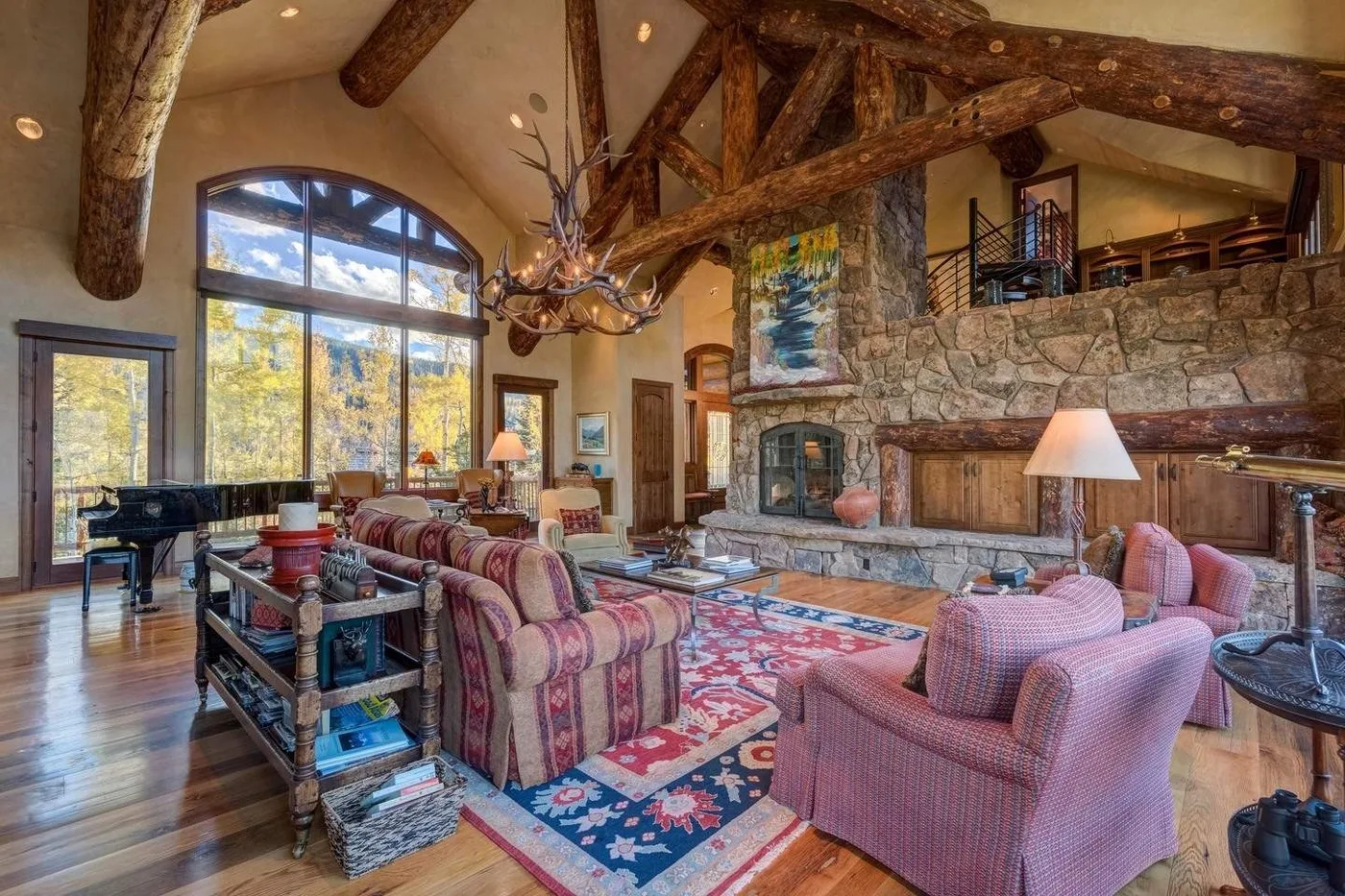A spacious living room has patterned seating around a stone fireplace beneath exposed timber beams.