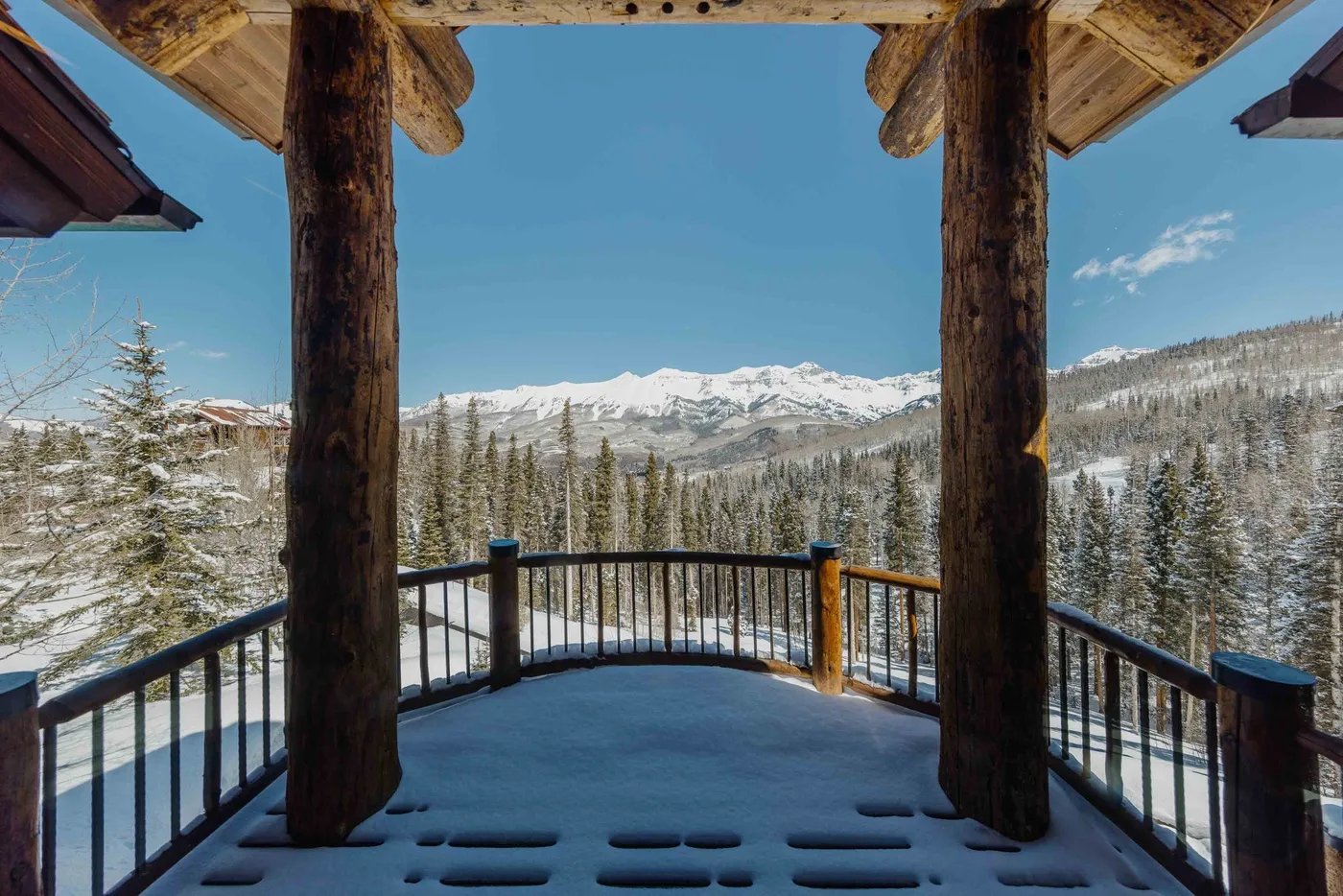 A snow-covered wooden deck overlooks a mountain range and evergreen trees.