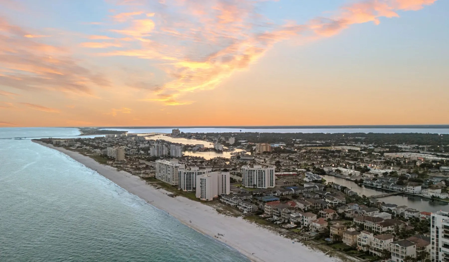 Vista aérea costera con una playa de arena y edificios bajo un cielo colorido al atardecer.