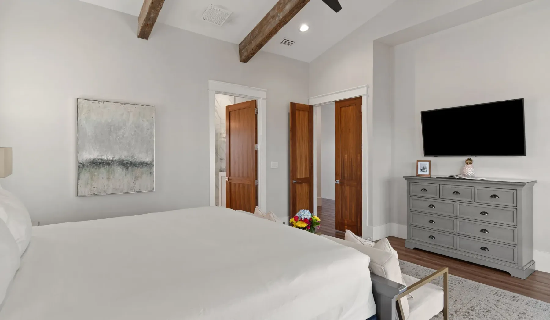 Un dormitorio luminoso con una cama, puertas de madera y una cómoda bajo un televisor en la pared.