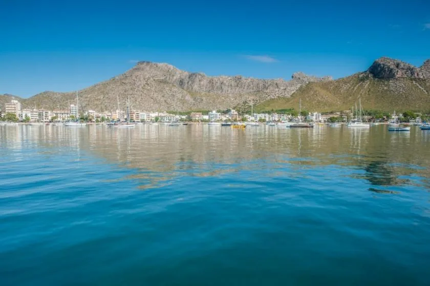 A coastal town sits along calm blue water beneath a rugged mountain range.