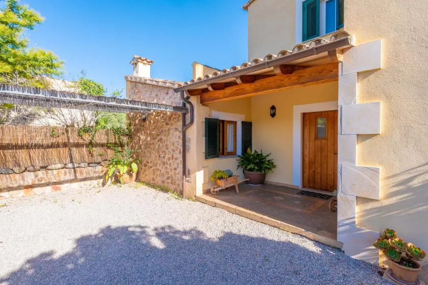 A house entrance opens onto a gravel courtyard with potted plants.
