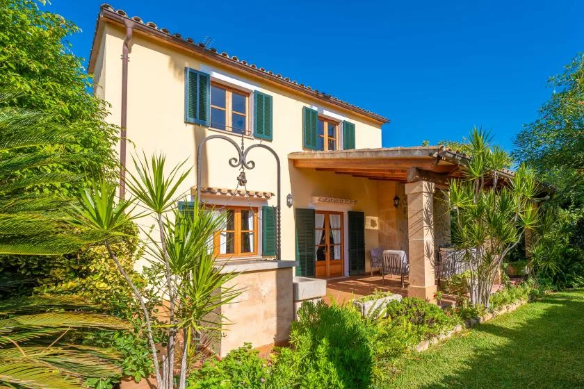 A cream-colored house with green shutters stands among garden plants under a clear blue sky.