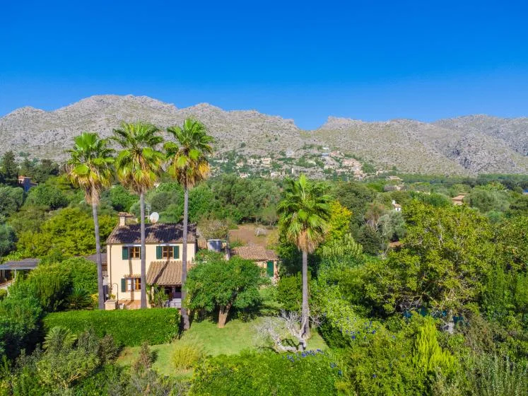A house and palm trees stand amid green gardens, with mountains in the background.